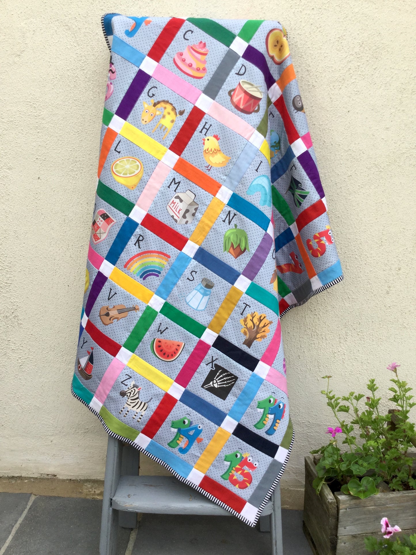Colourful sashing with cornerstones quilt with alphabet and illustrations draped over a chair outdoors.